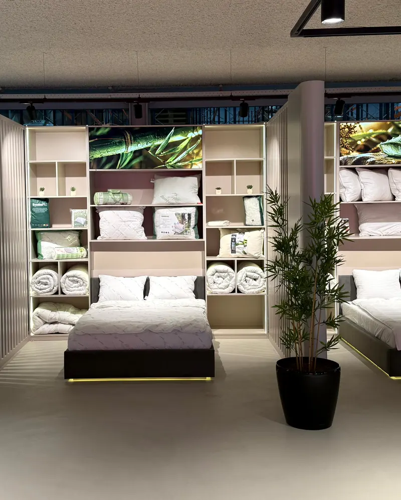 Der Showroom von HMG: ein gemachtes Bett zwischen Wandregalen mit Kissen und Bettdecken